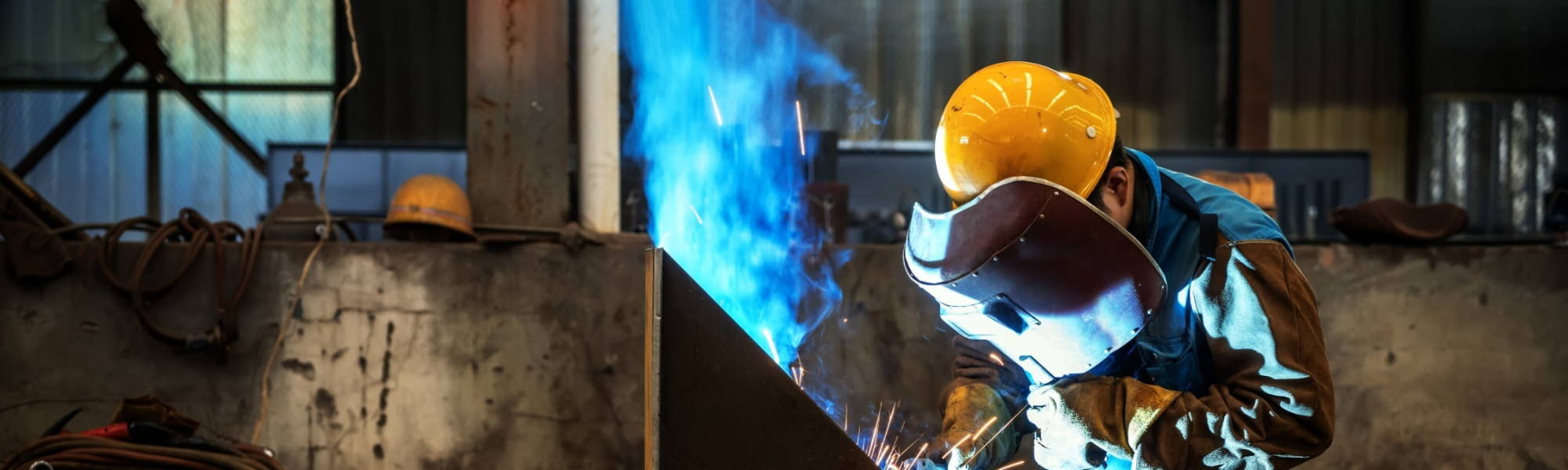 Man in yellow helmet welding metal with blue flame and smoke